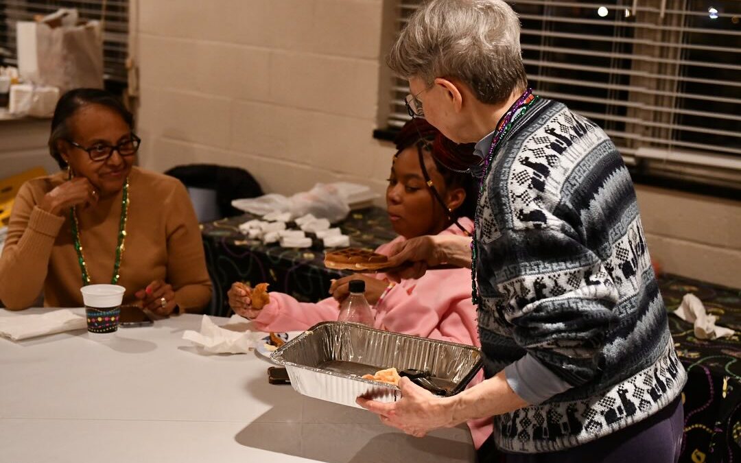 Three people at a table enjoying food together. One person serves from a tray while others eat and drink, creating a friendly atmosphere.