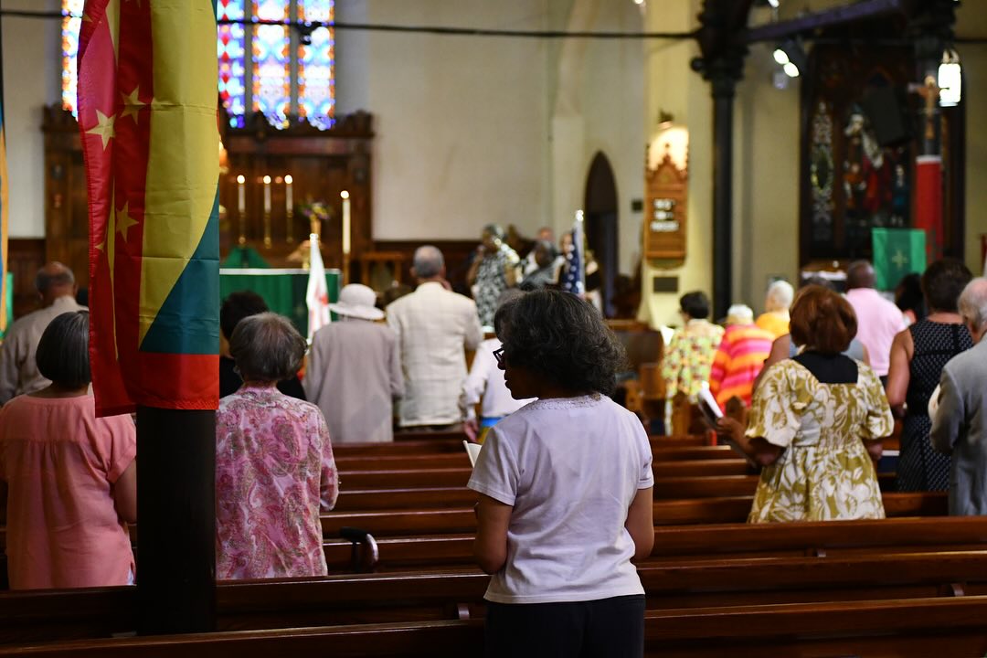 A church interior with stained glass, flag display, and people in pews facing the altar, participating in a religious service.