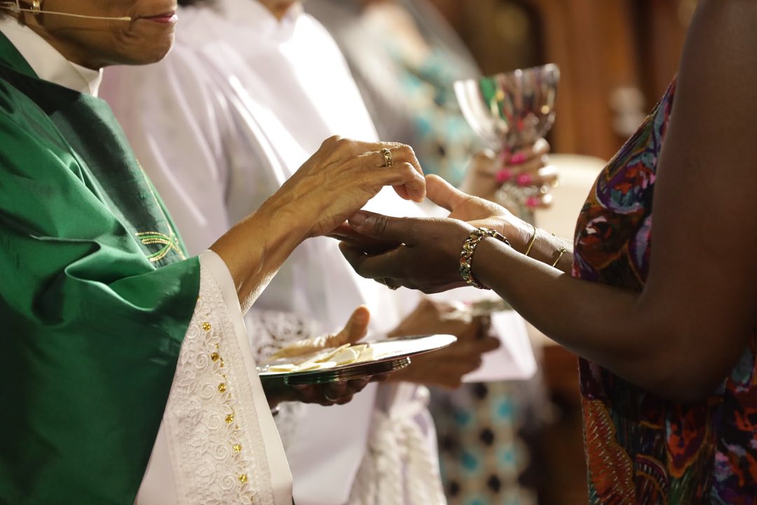 A person in religious attire distributes communion wafers to another person in a church setting, holding a chalice and plate.