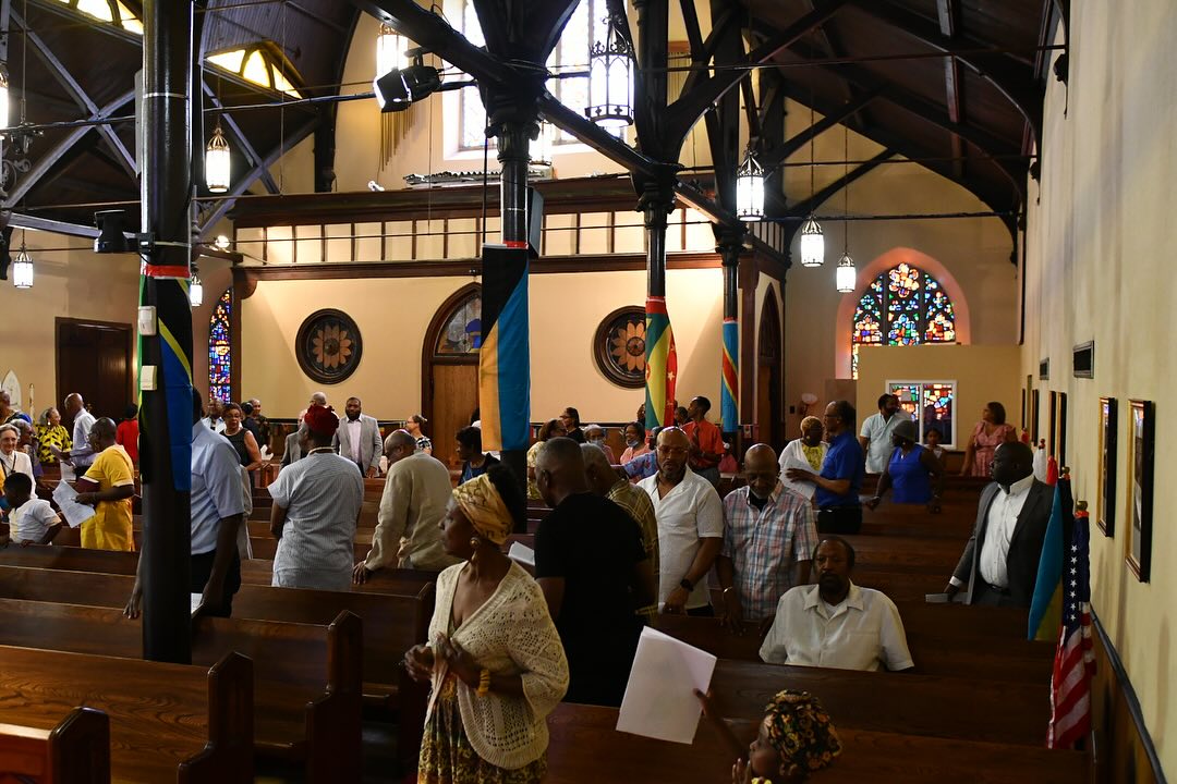 A group of people gathers inside a historic church, surrounded by stained glass windows and wooden pews, with various flags hanging from columns.